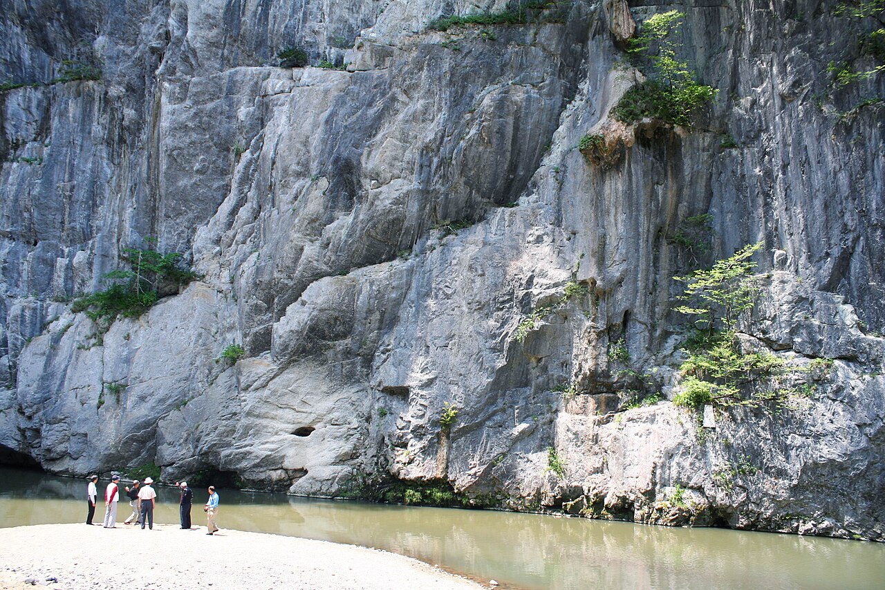 A huge grey limestone cliff face rising straight from the river at Geibikei, with a few visitors standing on the gravel bar at its foot