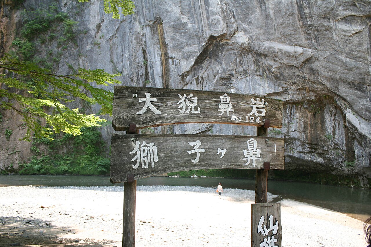 Wooden signposts naming Daigeibi-gan, 124 metres, and Shishigahana, beneath the overhanging limestone wall above a sandy river bank