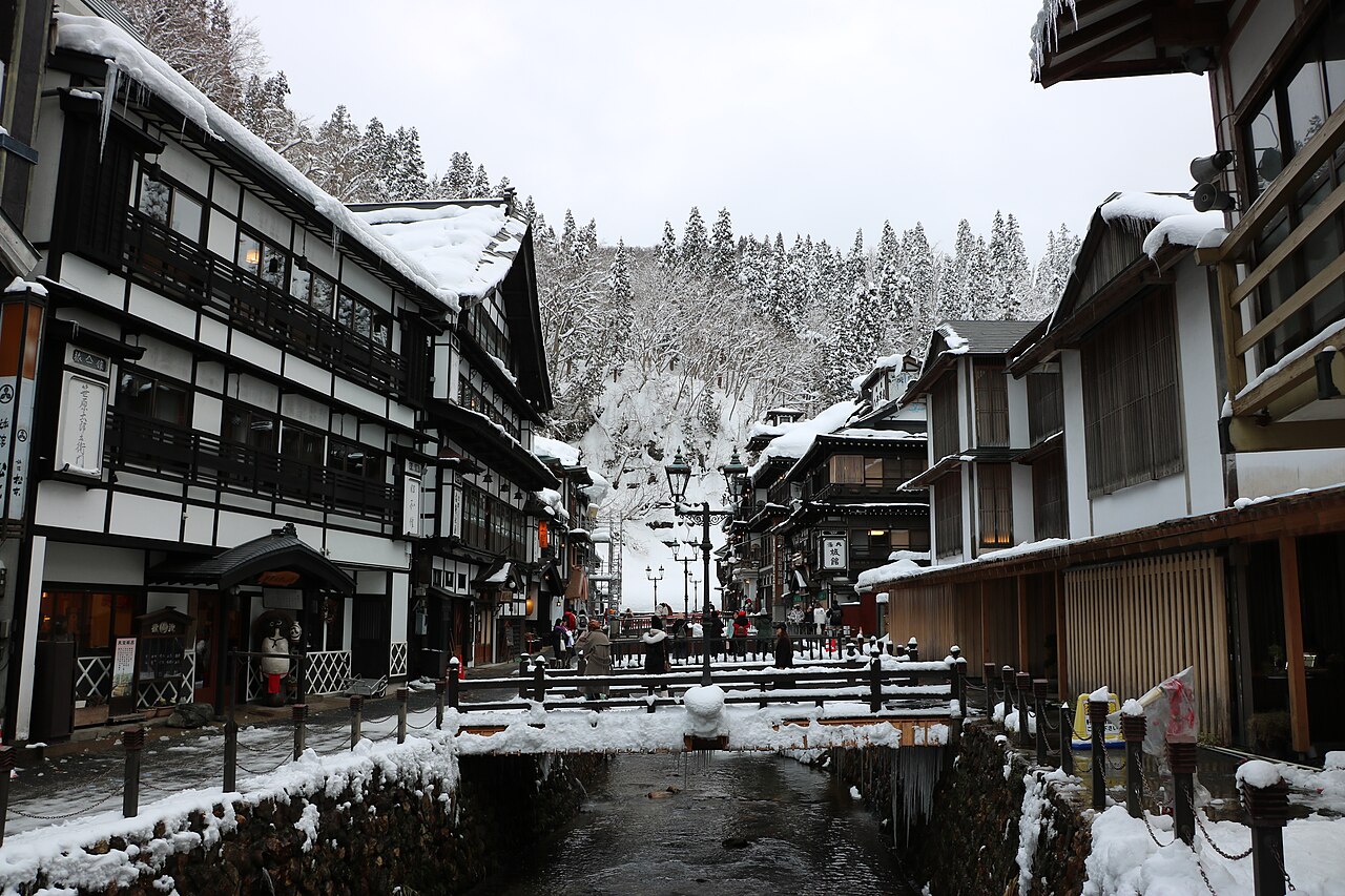 A snowy street in an onsen town with dark wooden multi-storey inns on both sides, a small bridge over a stream, and snow-covered trees on the hills behind.