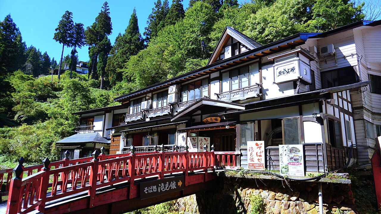 A red wooden bridge over a stream leading to a wooden building with a soba restaurant sign, with green forested hills behind.