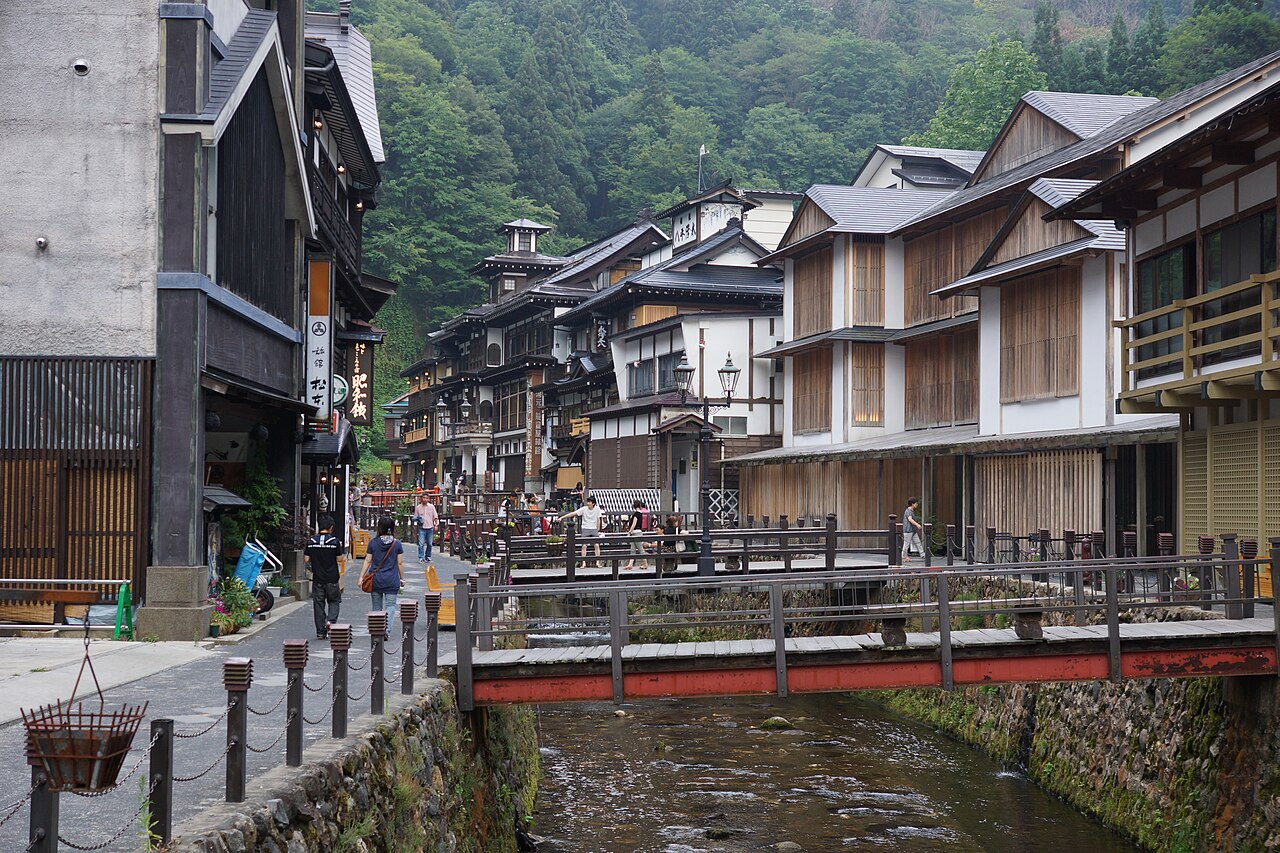 Traditional wooden inns beside a small river with a red bridge in the green mountains of an onsen town in summer.