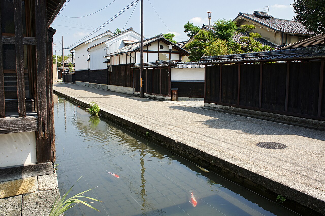 A narrow canal with two orange-and-white koi carp runs beside a paved lane lined with dark plank fences and white-walled storehouses under a blue sky.