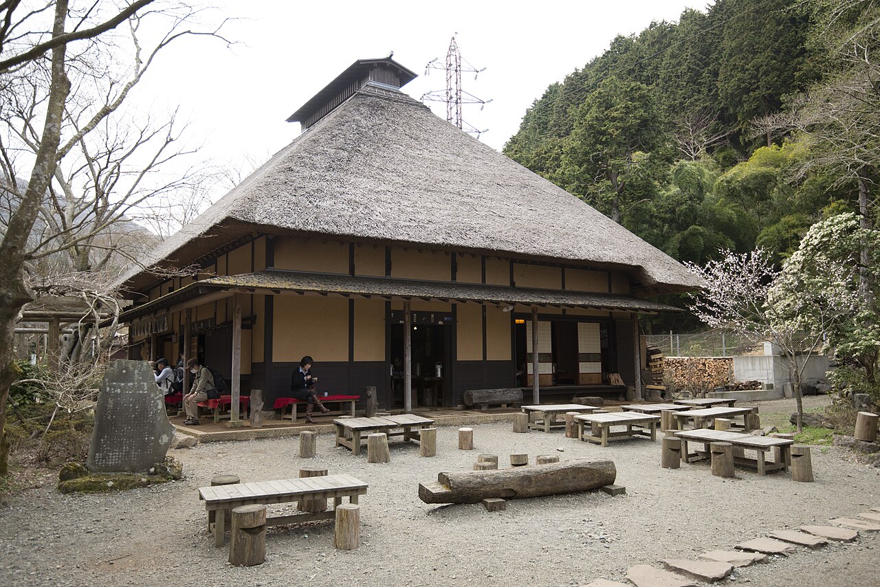 A large thatched-roof teahouse with low wooden benches and tables set out on the gravel in front, under bare trees and a wooded hillside