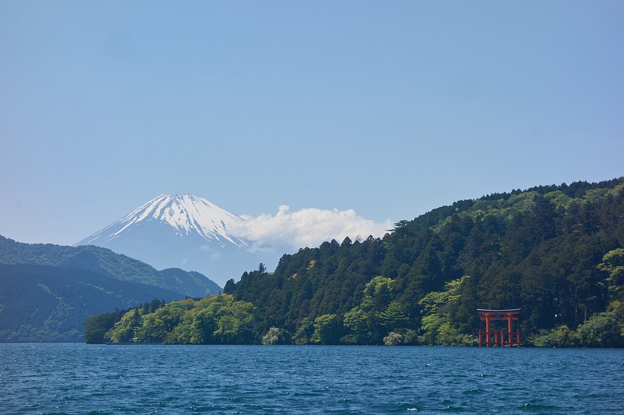Snow-capped Mount Fuji rising beyond the wooded shore of Lake Ashi, with a red torii standing at the water's edge on the right