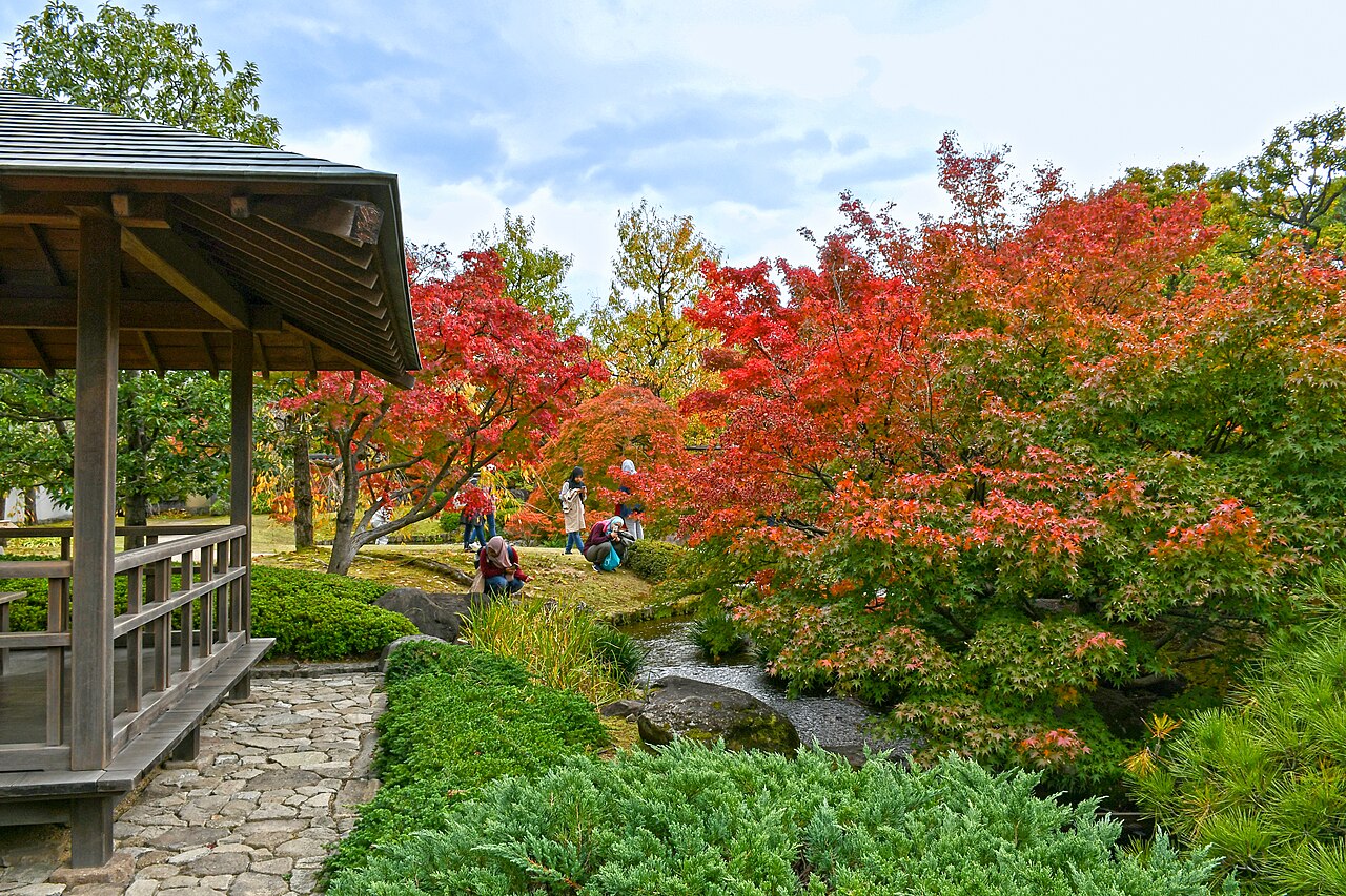 A wooden pavilion with a railing beside a stone path at left, looking over a small stream, clipped shrubs and red-leaved trees, with a few visitors in the distance.