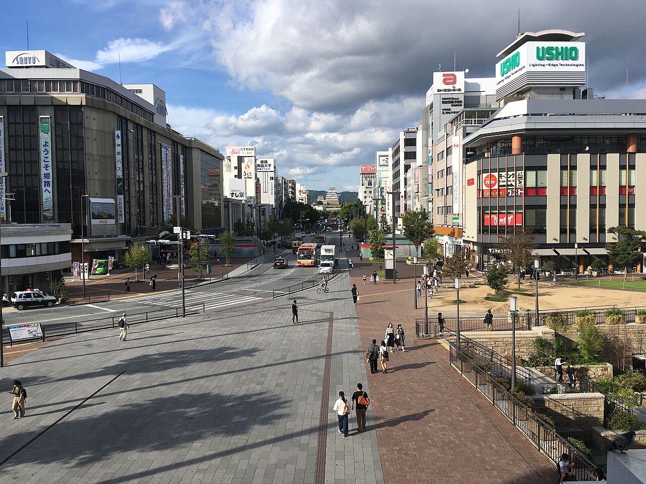 Looking down from above onto a wide paved plaza and a broad avenue between multi-storey buildings, with buses on the road and the white castle keep small in the distance.