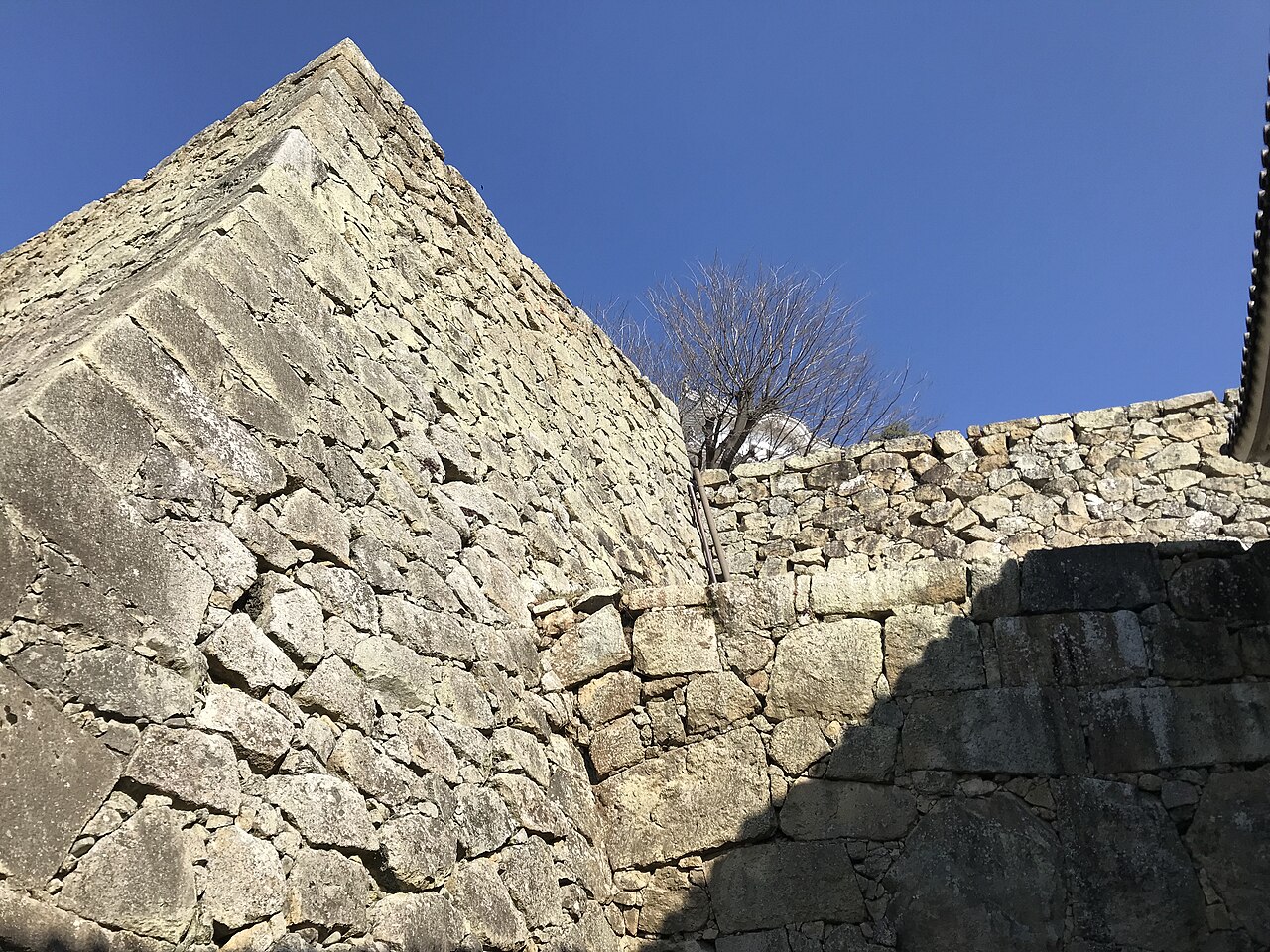 A tall stone wall of fitted rough stones with a sharp angled corner, and a lower stone wall and bare branches against a clear blue sky.