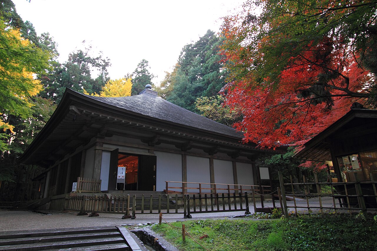 A wooden hall with a dark roof and white wall panels beside bright red and yellow autumn maple leaves, with stone steps at lower left.