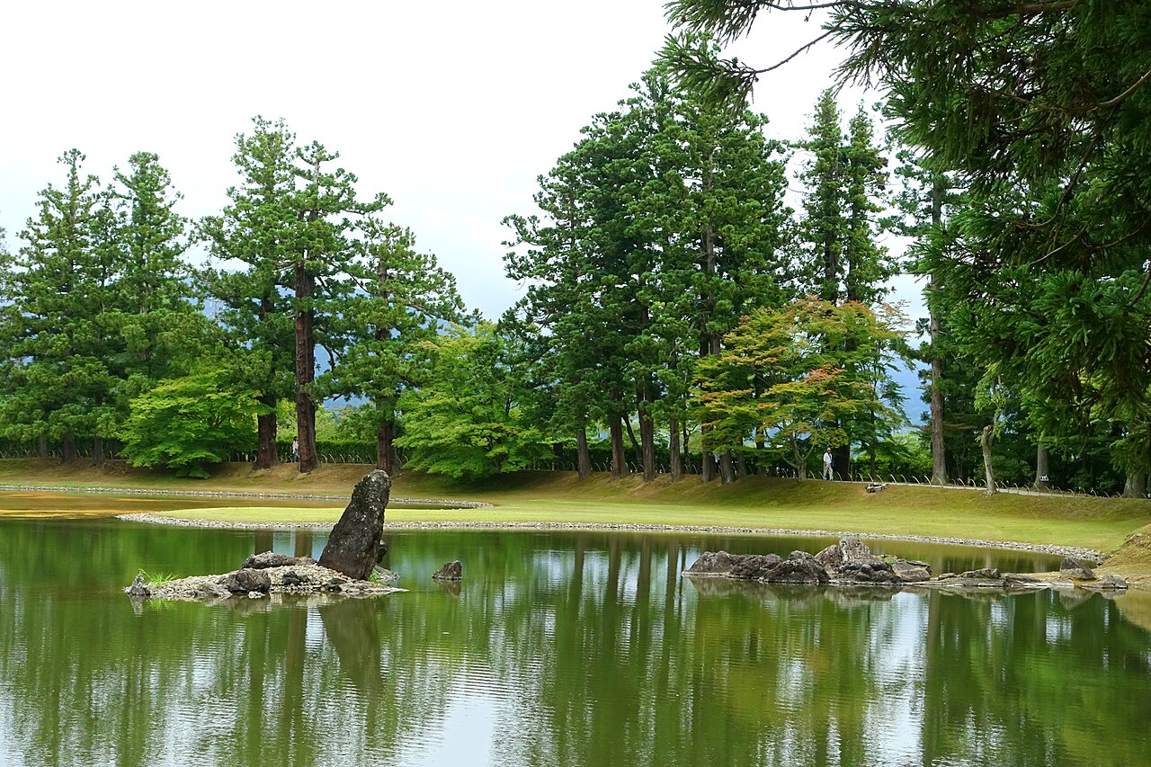 A still pond with a standing rock and small stone islets in the foreground, a grassy bank and a line of tall conifers reflected in the water.