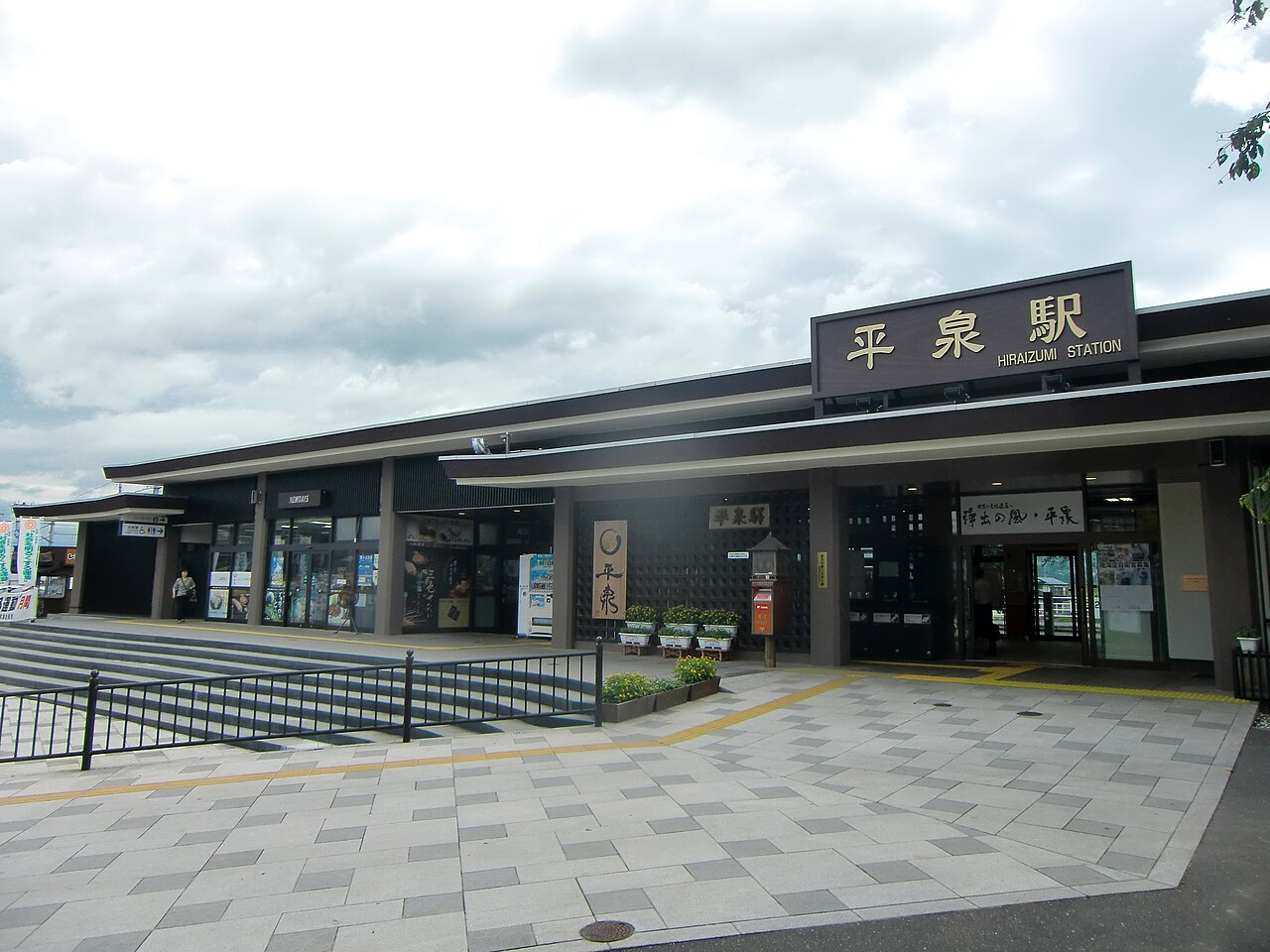Front of Hiraizumi Station, a low dark-roofed building with the station name sign above the entrance, a paved forecourt and a wide staircase at left.