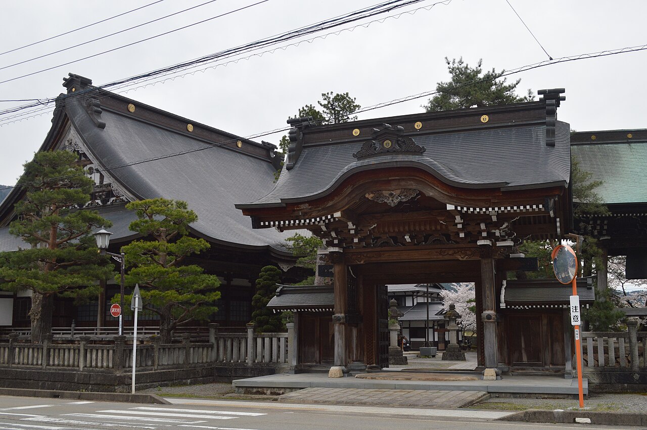 The carved wooden gate of Honkoji temple in Hida Furukawa, with the large grey-tiled main hall and pine trees behind it