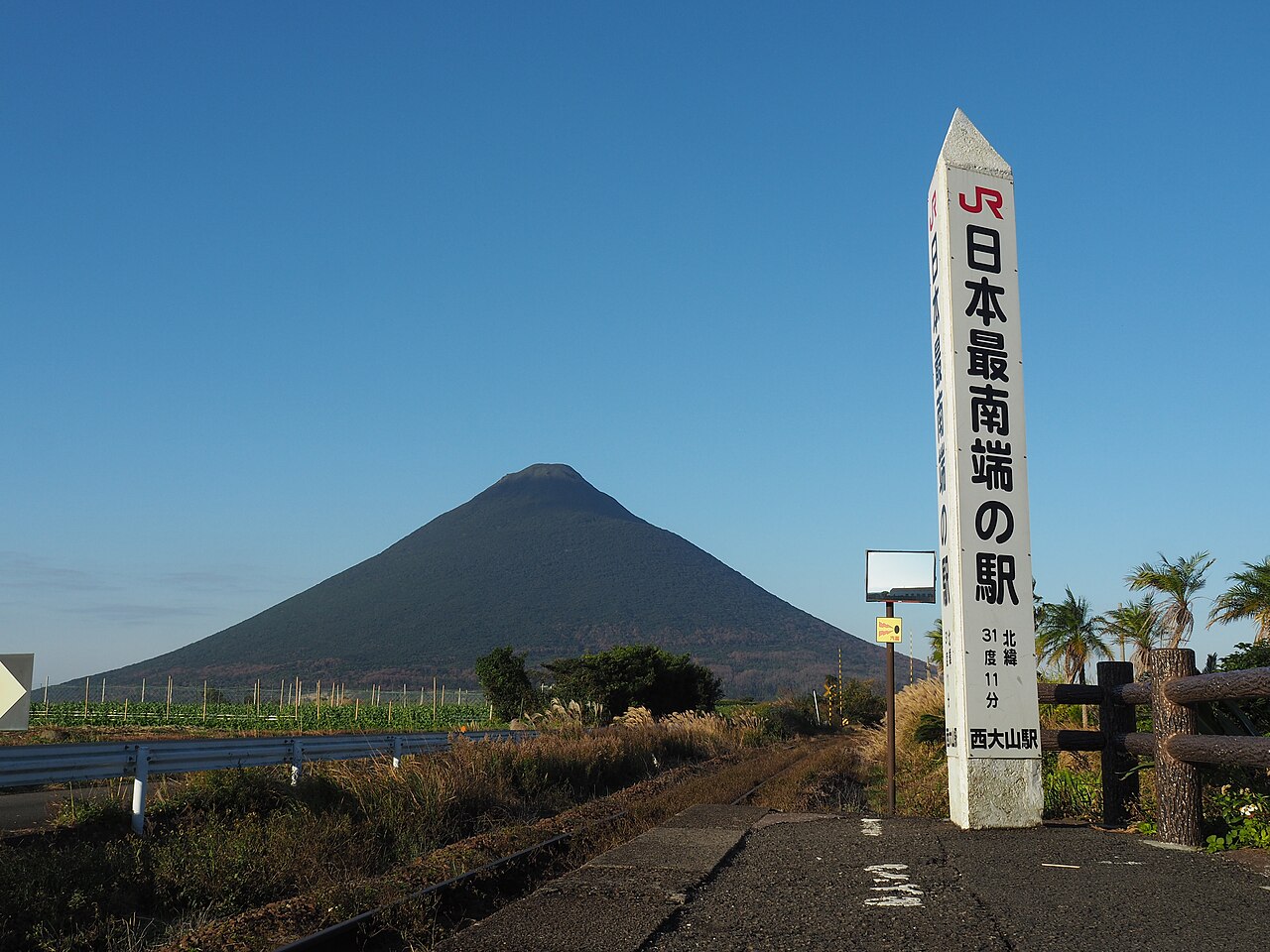 The white marker reading Japan's southernmost station on the platform at JR Nishi-Oyama Station, with Kaimondake behind