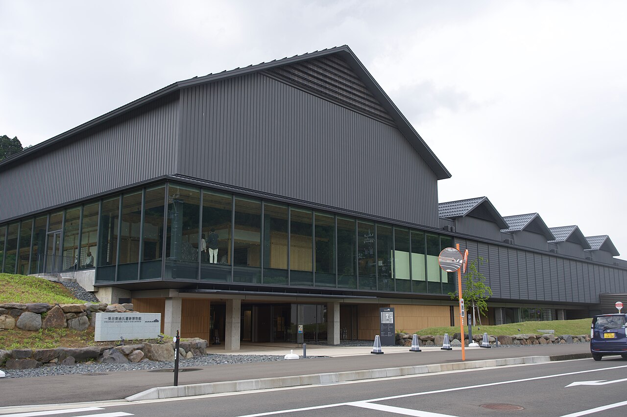 The new building of the Ichijodani Asakura Family Site Museum, a dark gabled structure with a glass-fronted ground floor and a stone sign at the roadside