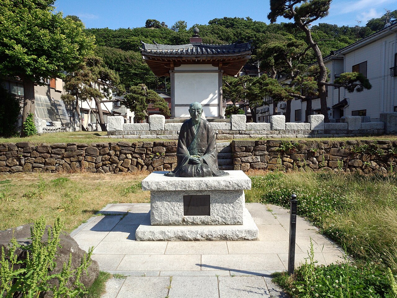 Seated statue of Ryokan on a stone pedestal at his birthplace site in Izumozaki, with a small tiled-roof hall behind.