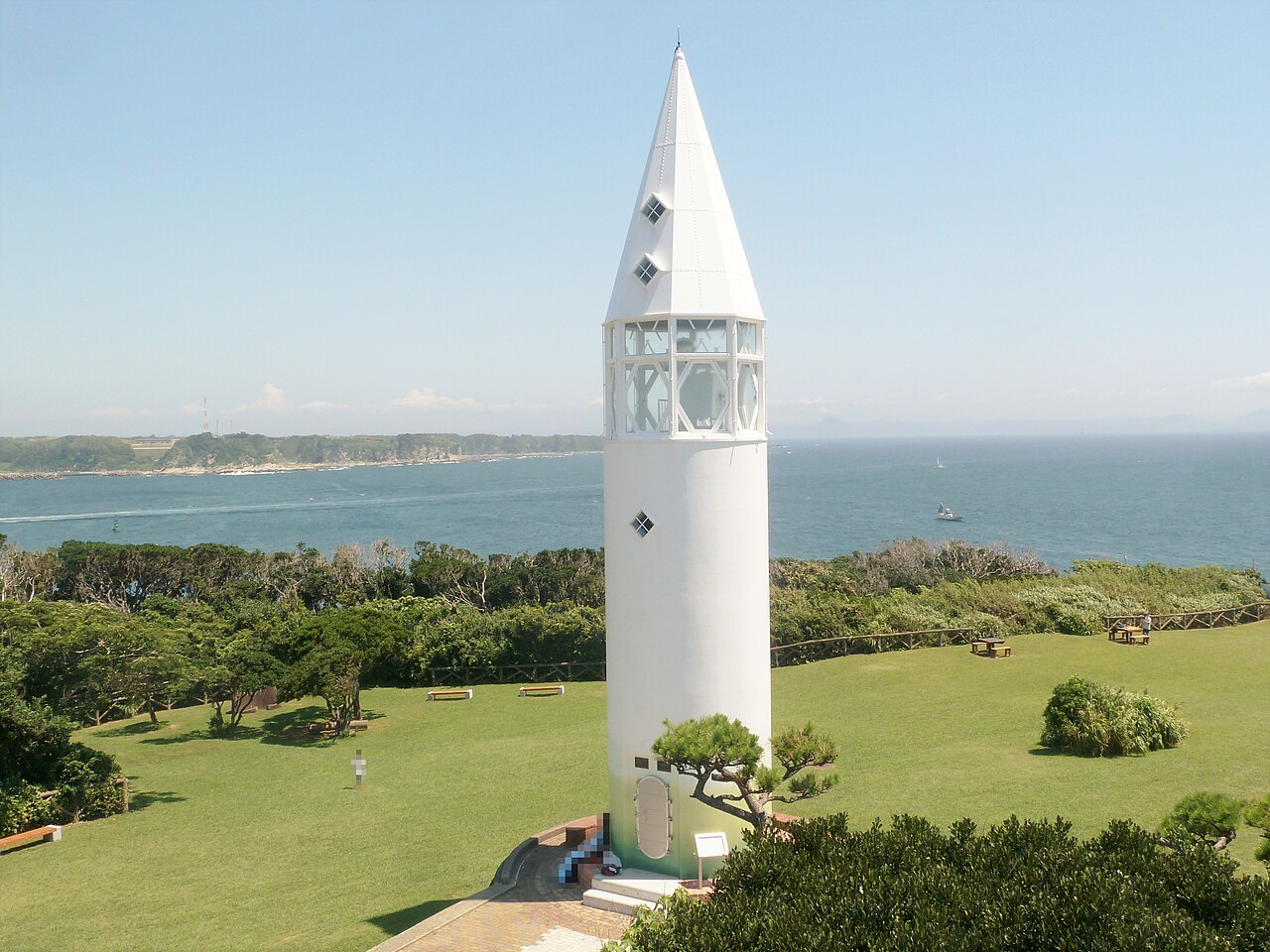 A slim white conical Awasaki Lighthouse with small diamond-shaped windows on a green lawn with the sea and a distant shoreline behind