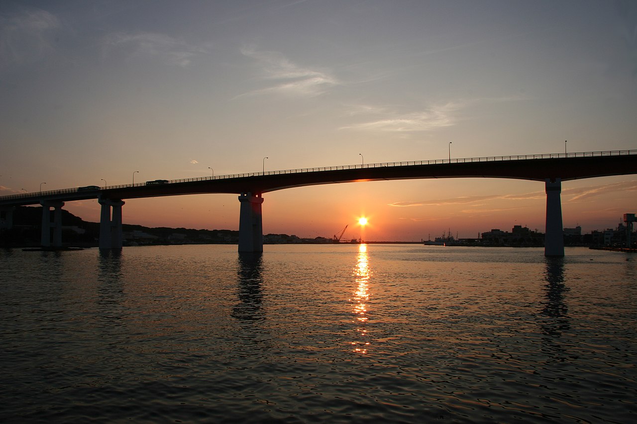 A low orange sun on the horizon behind the dark spans and piers of Jogashima Bridge, reflected in calm water