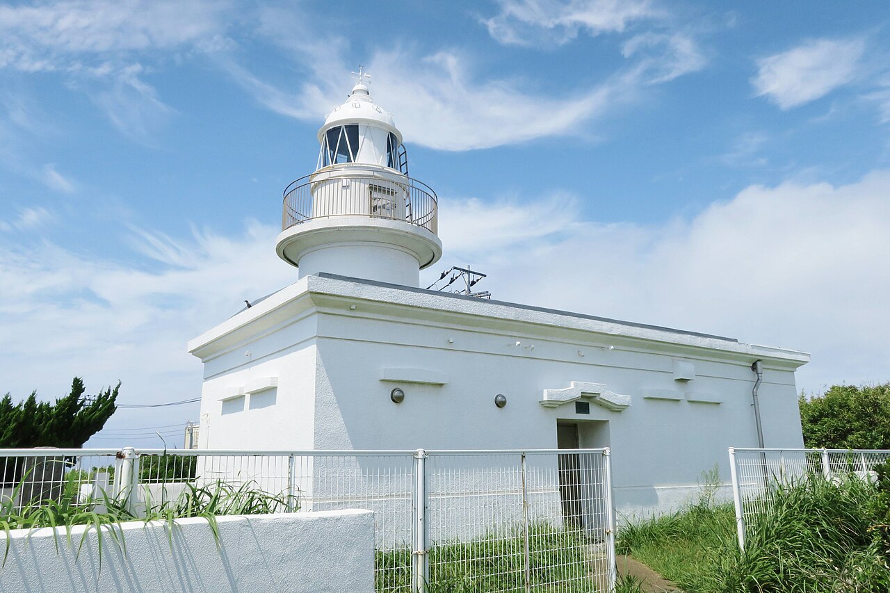 A white Jogashima Lighthouse tower with a lantern room on a low white building under a blue sky with wispy clouds, seen from a grassy path