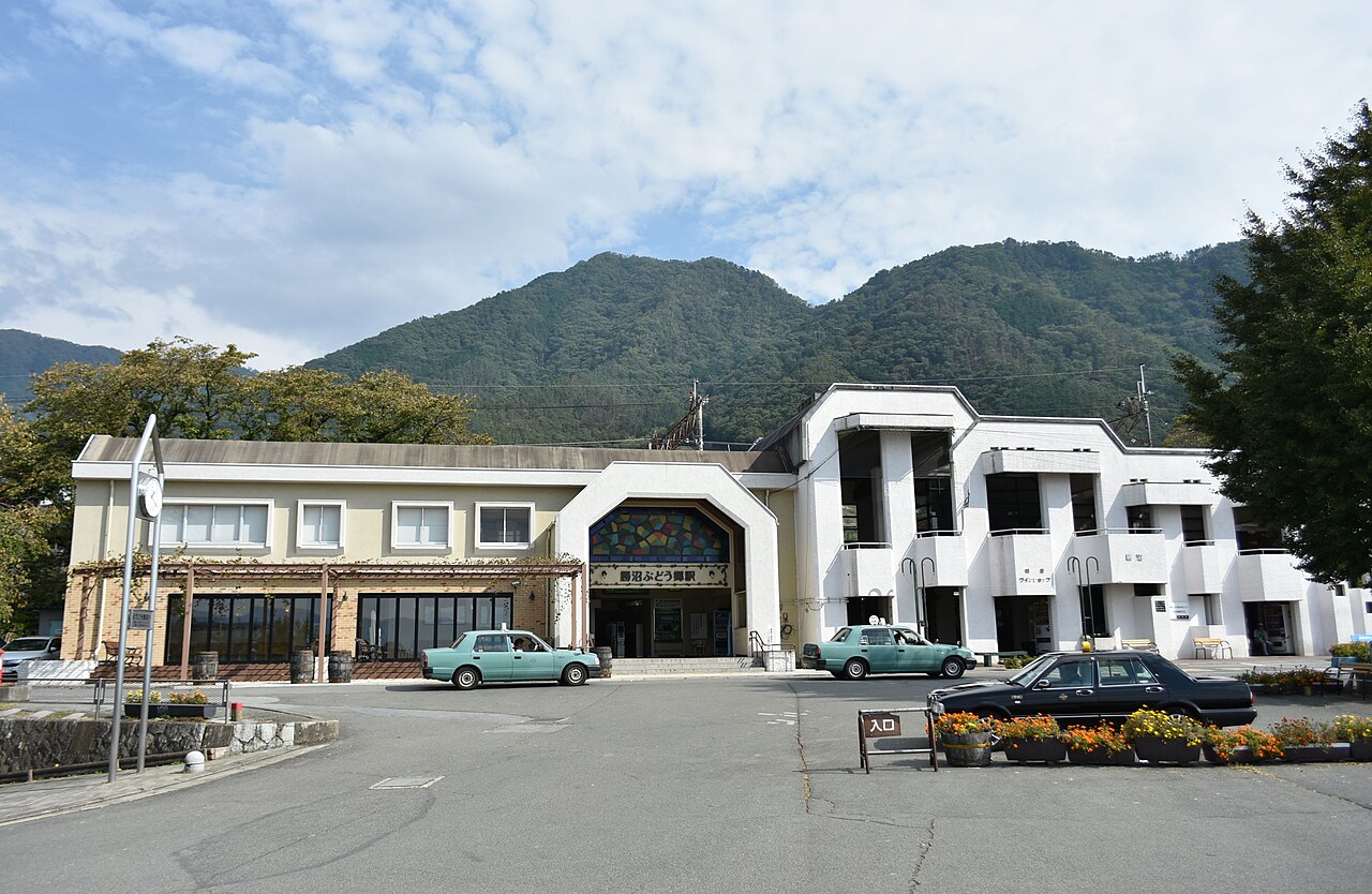 The white Katsunuma-budokyo station building with taxis waiting in front and green mountains behind.
