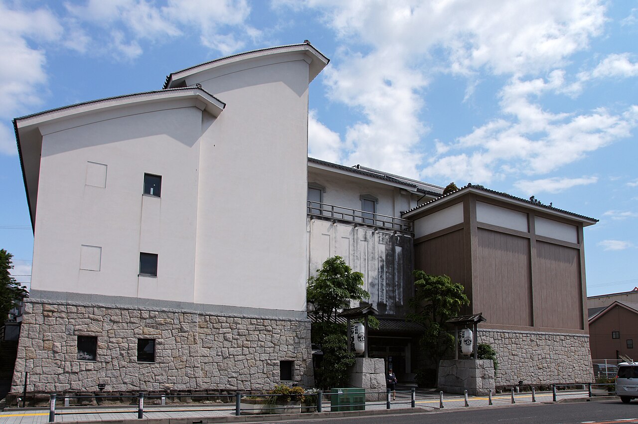 The exterior of the Kishiwada Danjiri Kaikan, a cream-walled building with curved eaves and a stone-faced base, with paper lanterns at the entrance under a blue sky with clouds.