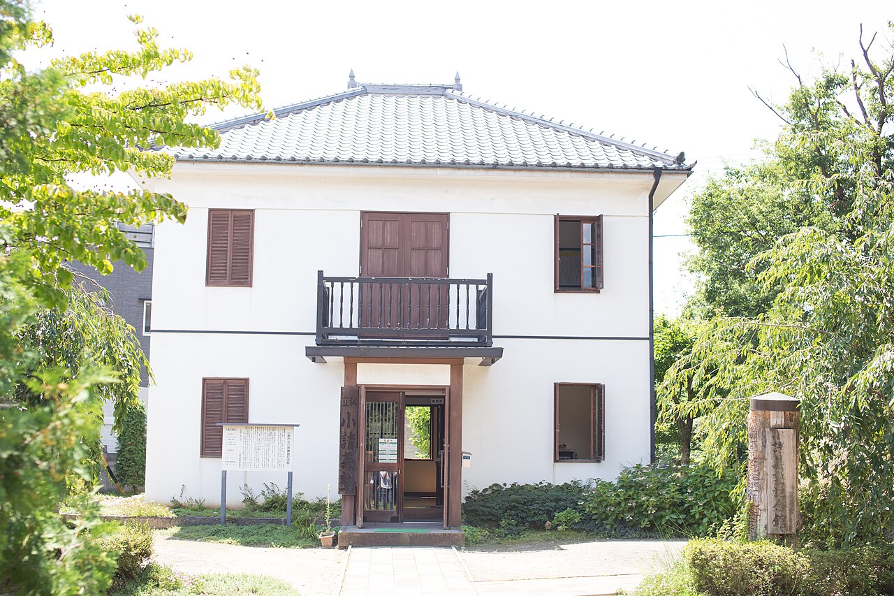 The two-storey white-walled Komoro Gijuku Memorial Hall with a wooden balcony and entrance porch, surrounded by greenery.