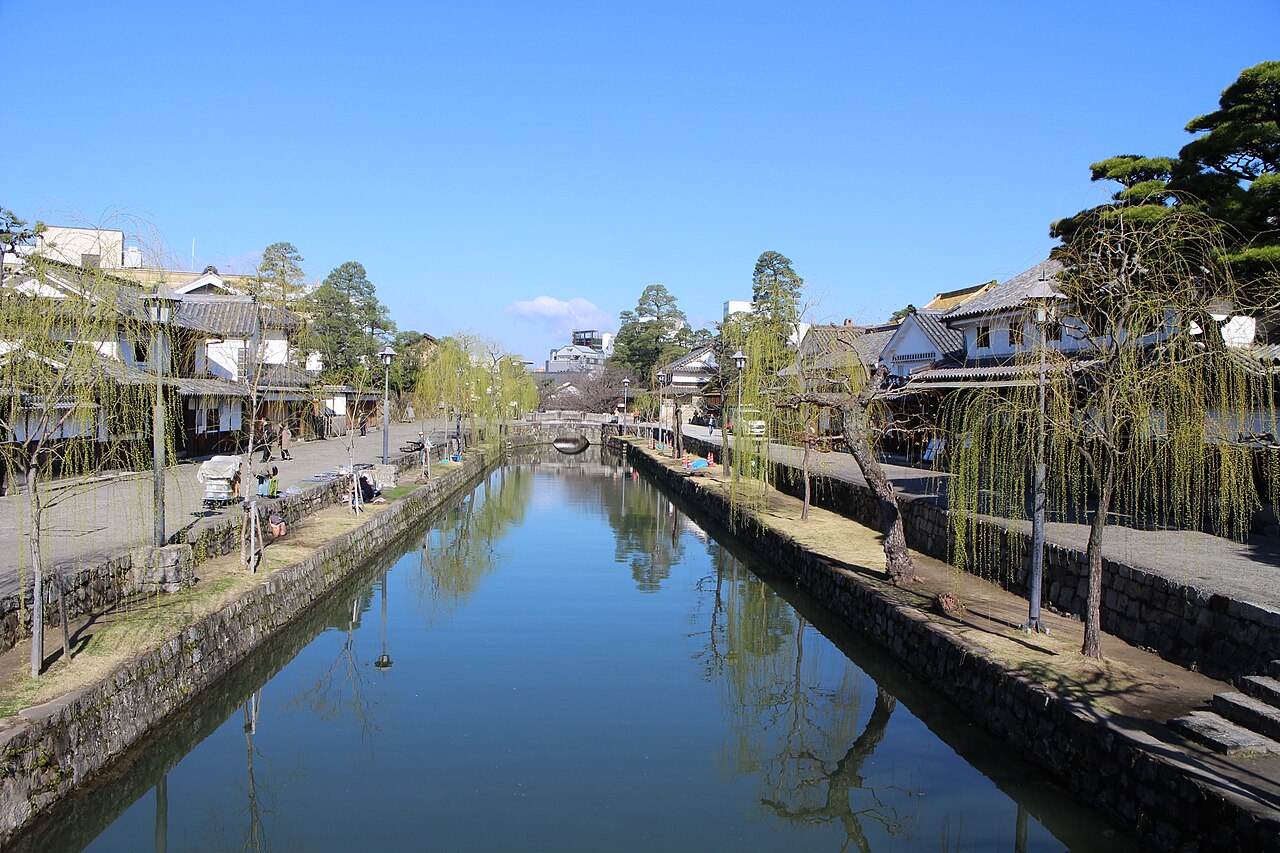 A straight canal reflecting a blue sky, lined with stone embankments, weeping willows and low white-walled buildings on both banks.