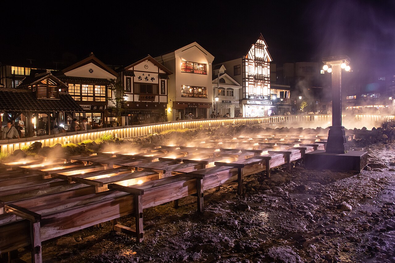 The wooden flumes of the Yubatake lit warmly at night with steam rising, a tall street lamp on the right and lit wooden buildings of the old town behind.