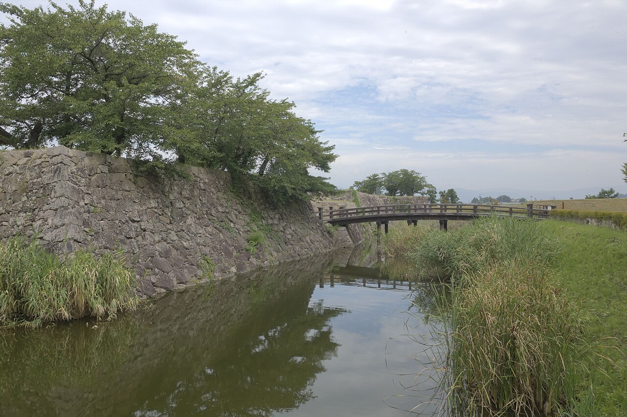 The water-filled moat of Matsushiro Castle, with a mossy stone wall on the left, reeds on the right and a low wooden bridge in the distance.