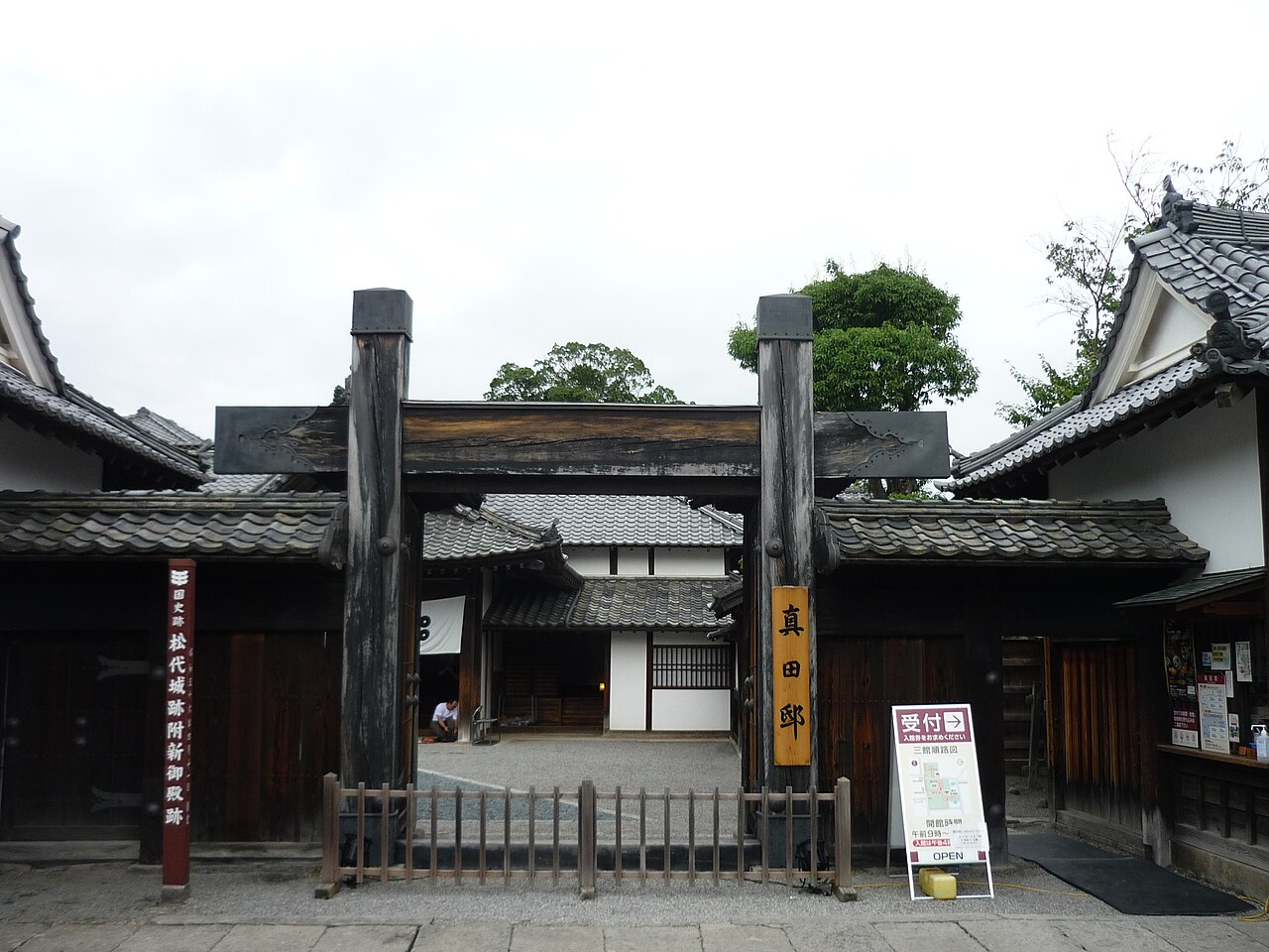 The dark wooden entrance gate of the former Sanada Residence in Matsushiro, with tiled roofs and garden trees behind it.
