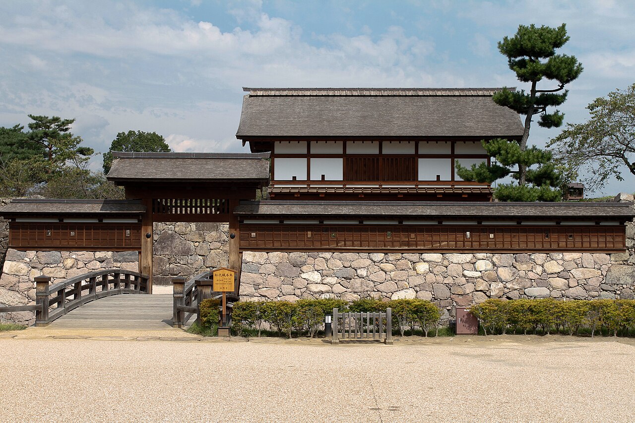 The reconstructed Taikomon gate of Matsushiro Castle, a white-walled wooden gatehouse on a stone base with a small bridge and a pine tree under a clear sky.