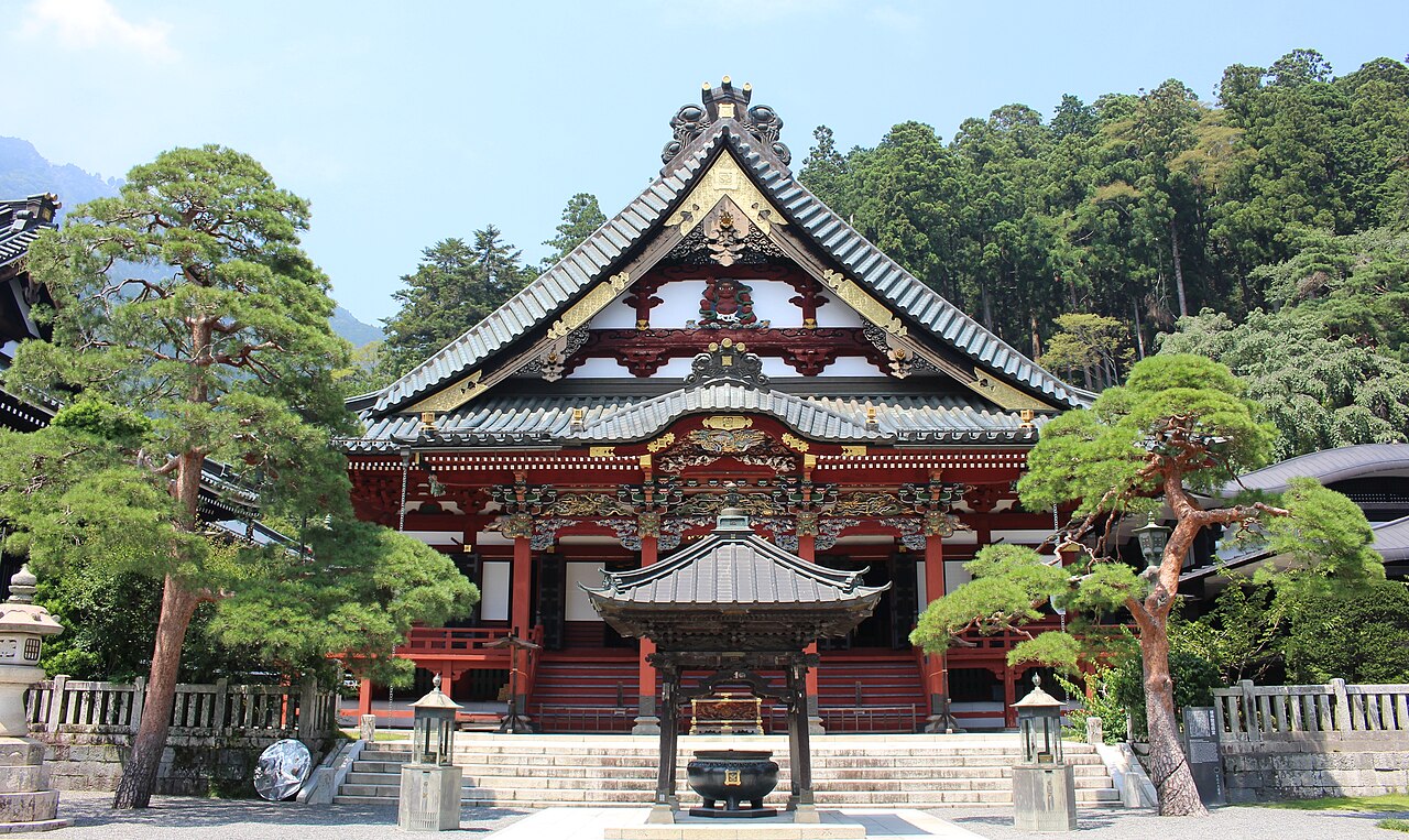 A red-painted hall with a carved gable, pine trees and an incense burner at Kuon-ji
