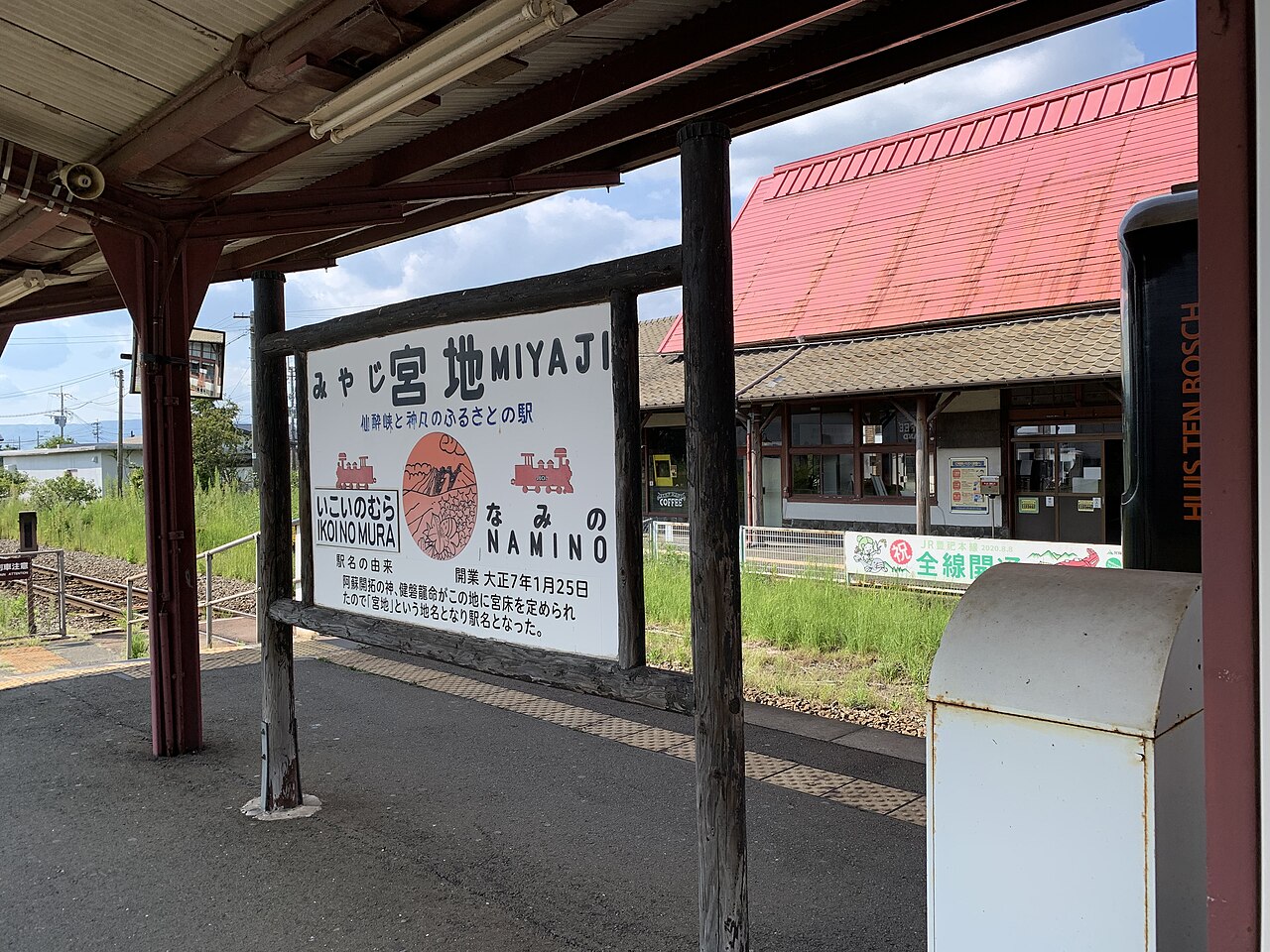A station nameplate reading Miyaji on a wooden-framed board on the platform, with a red-roofed station building and a rail line behind it.