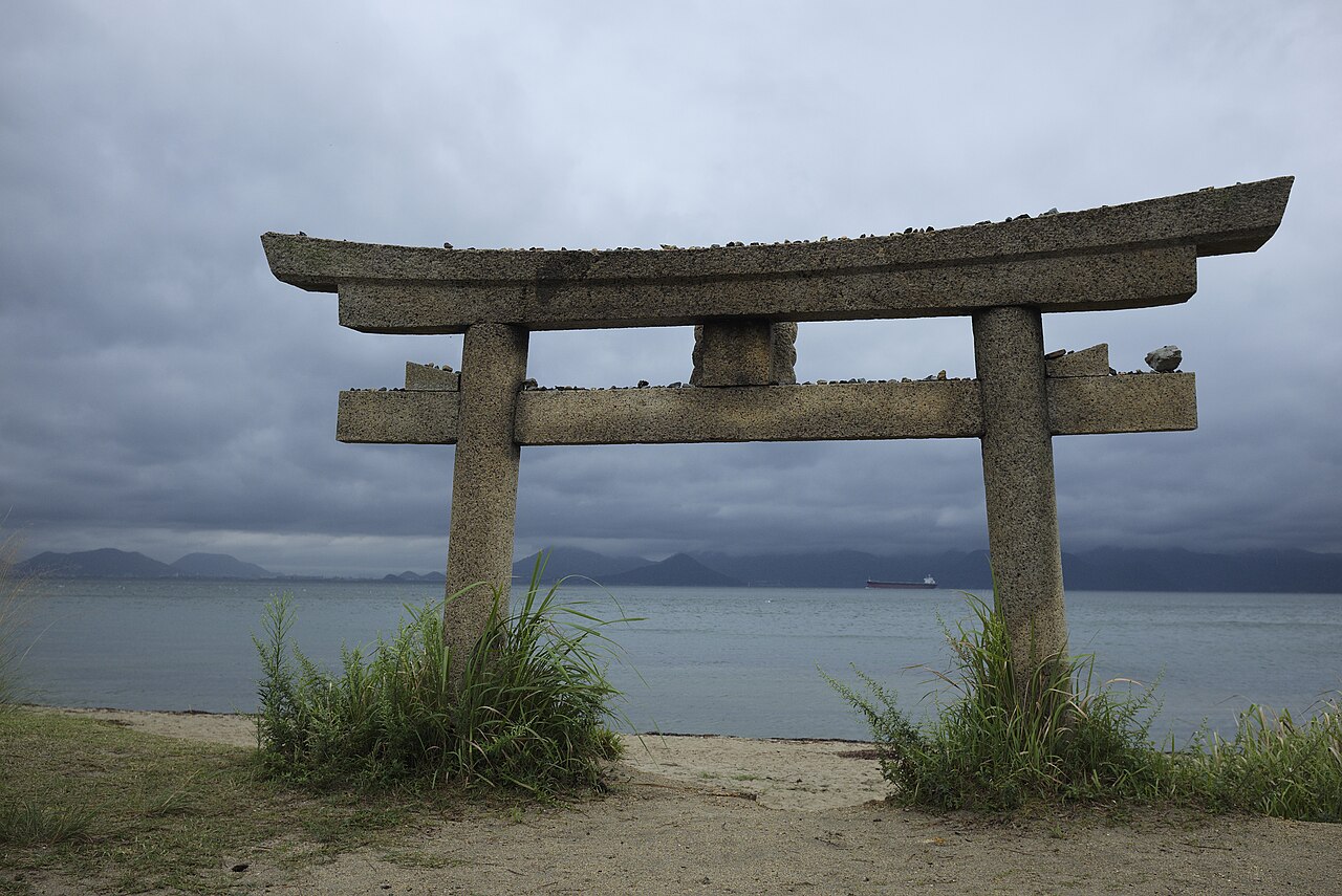 A stone torii half buried in sand on the shore at Kotohikichi beach on Naoshima, with the Seto Inland Sea beyond
