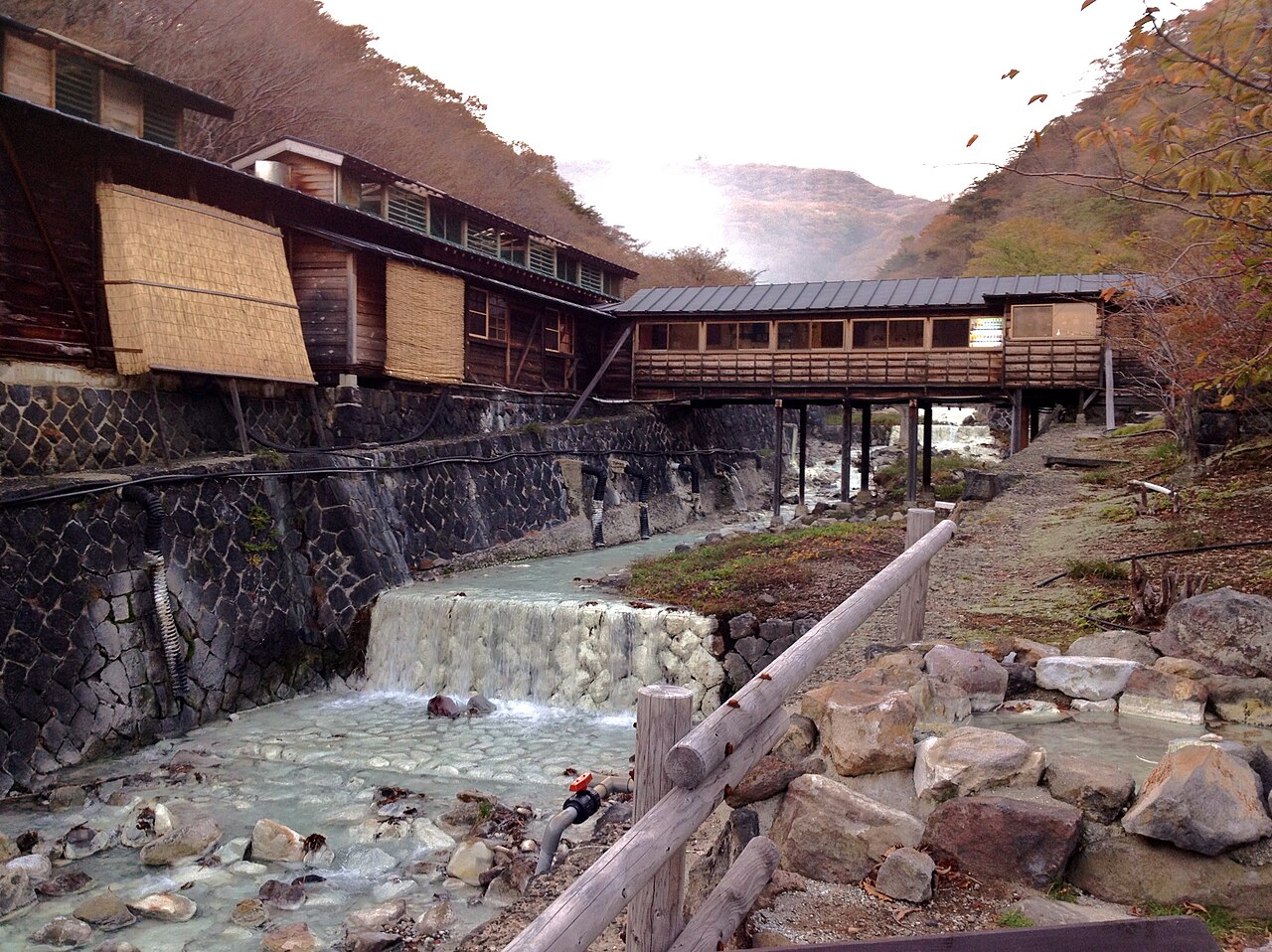 Wooden bathhouse buildings and a covered walkway above a clear stream at Shika-no-yu in Nasu Yumoto