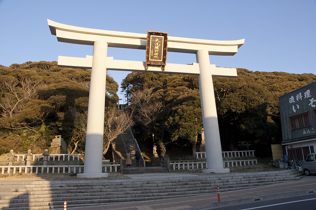 A tall white stone torii with a framed name plaque stands at the foot of a flight of steps, with dense pine and evergreen trees behind it