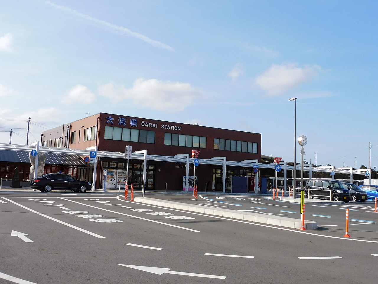 The low Oarai Station building with its sign above the entrance, seen from the wide forecourt and road on a clear blue day