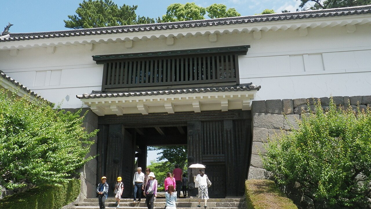 The black timber Tokiwagi Gate of Odawara Castle with white plaster walls, visitors climbing the stone steps under a clear sky, with greenery around.