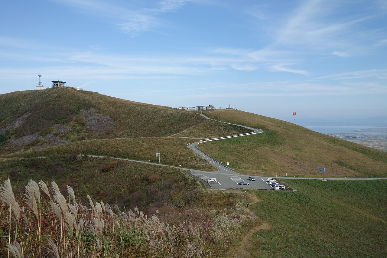 A winding road climbs the grassy slopes of Mount Kanpu on the Oga Peninsula, with a lookout building on the summit and a paraglider in the sky.