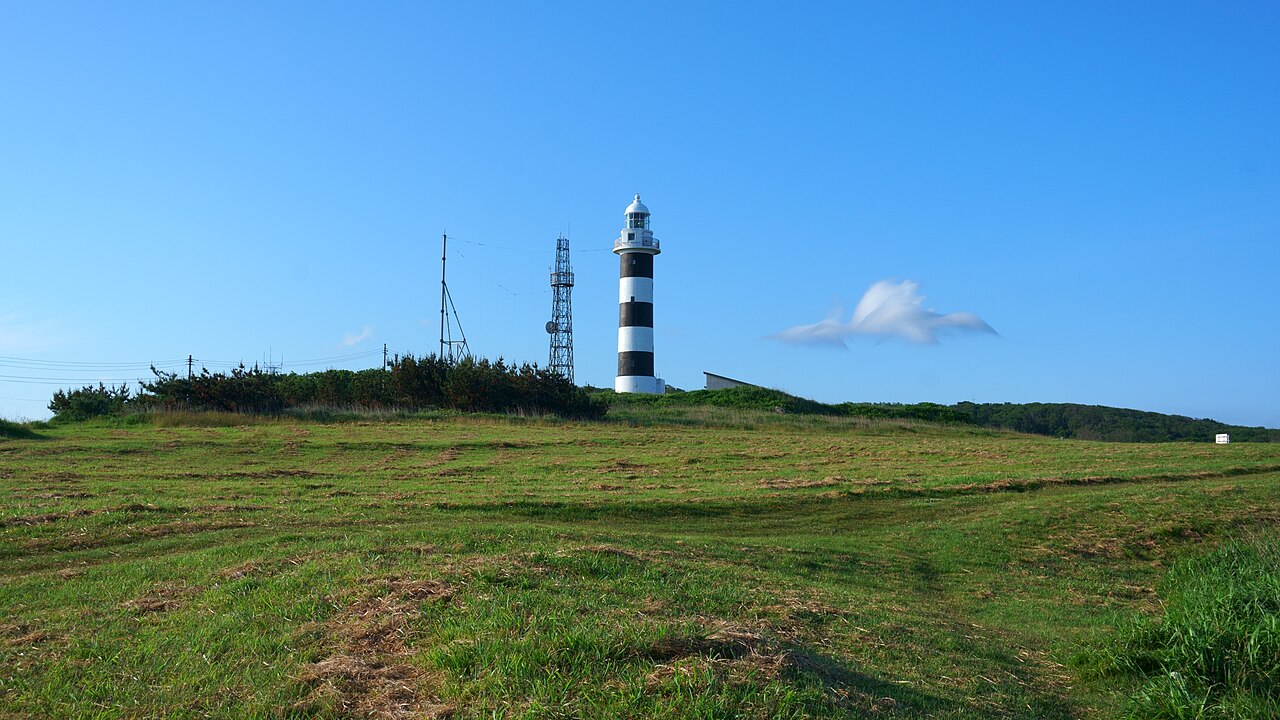 The black-and-white striped Nyudozaki Lighthouse standing above a mown grass field under a clear blue sky on the Oga Peninsula.