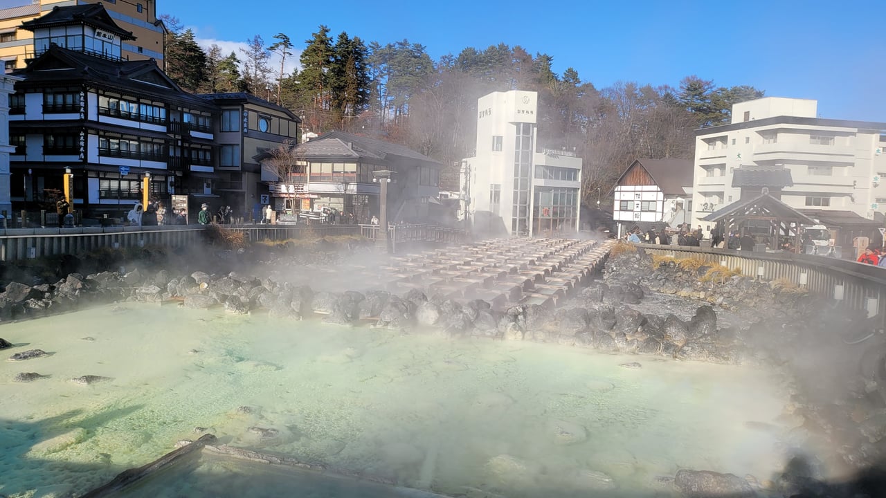 A pale turquoise hot-spring pool releasing thick steam among black rocks, with traditional wooden buildings on the hillside behind.