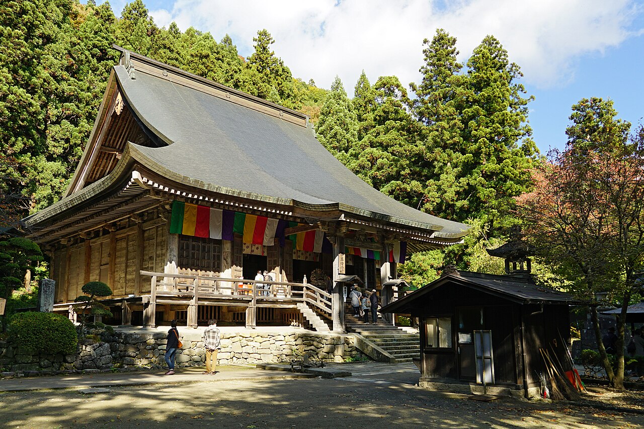 Risshaku-ji's Konpon Chudo main hall at Yamadera, its wide grey roof hung with five-colour cloth under tall cedars on a sunny autumn day