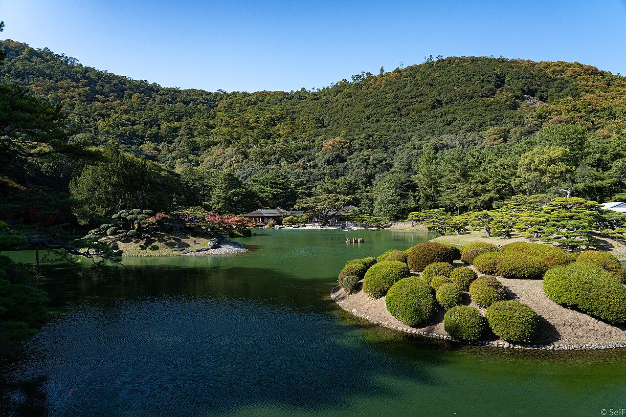 A pond at Ritsurin Garden with rounded clipped shrubs on the shore, a teahouse across the water and Mount Shiun rising behind