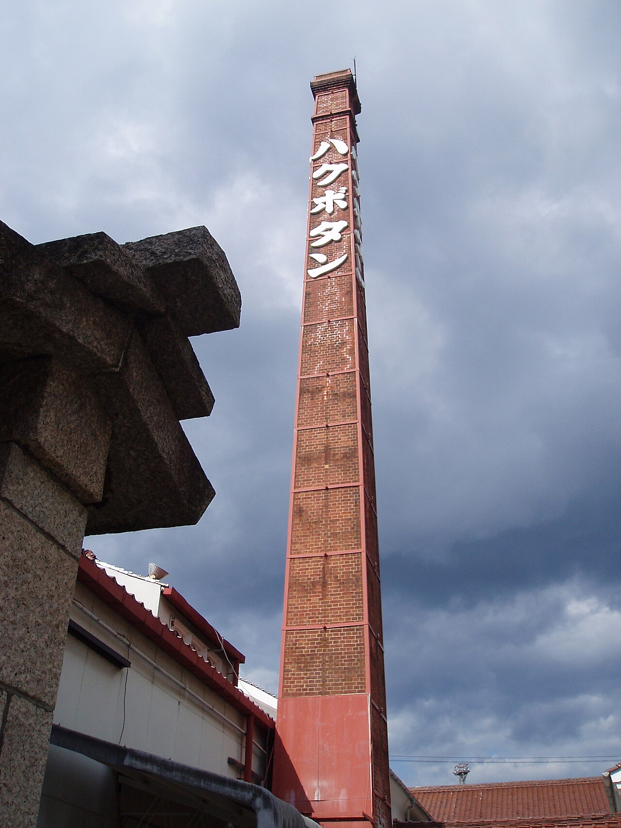 A tall red-brick brewery chimney with vertical katakana lettering seen from below, beside a stone lantern, against a cloudy sky.
