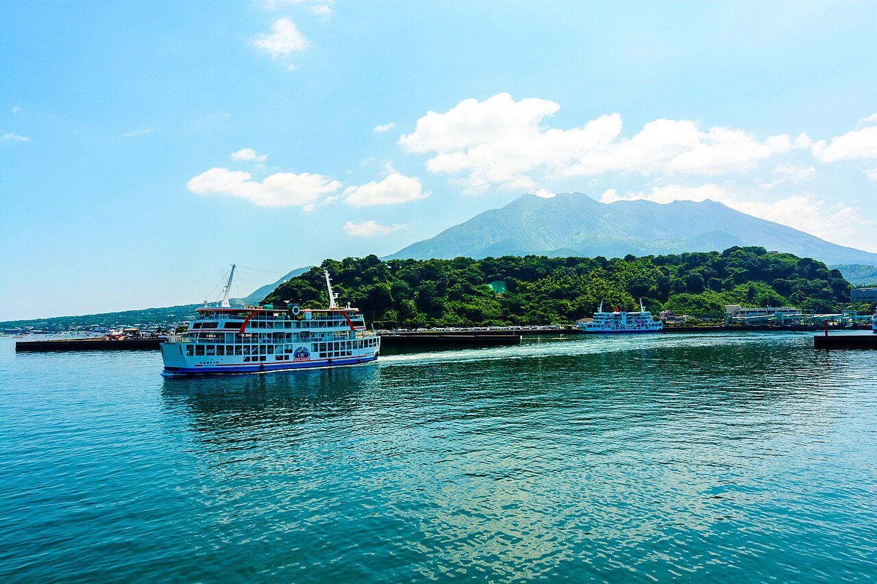 A white ferry on blue harbour water with the green slopes of Sakurajima rising behind a wooded shoreline