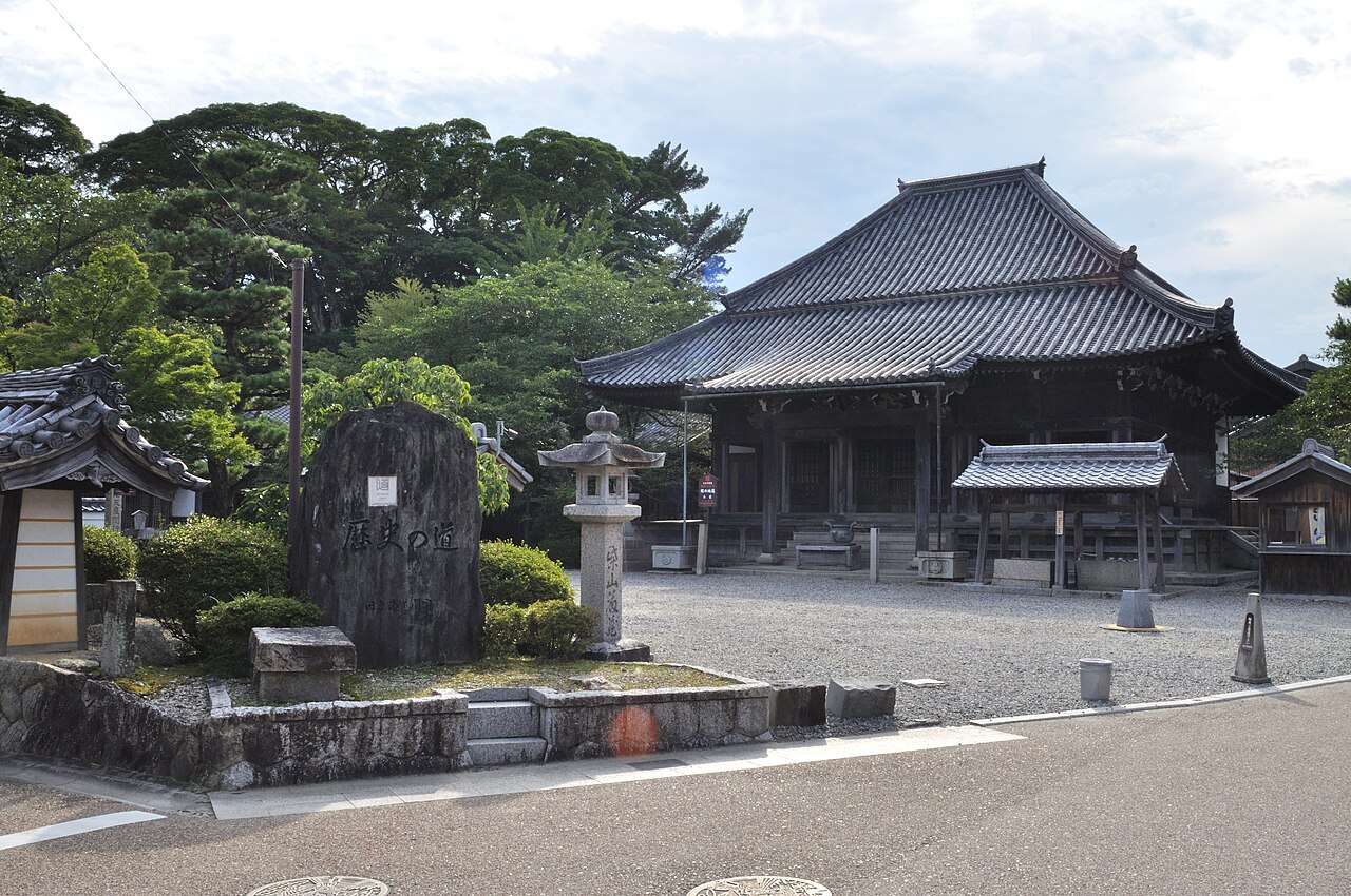 The tiled main hall of Seki Jizo-in behind a carved stone monument and a stone lantern in a gravel courtyard, with large trees beyond.