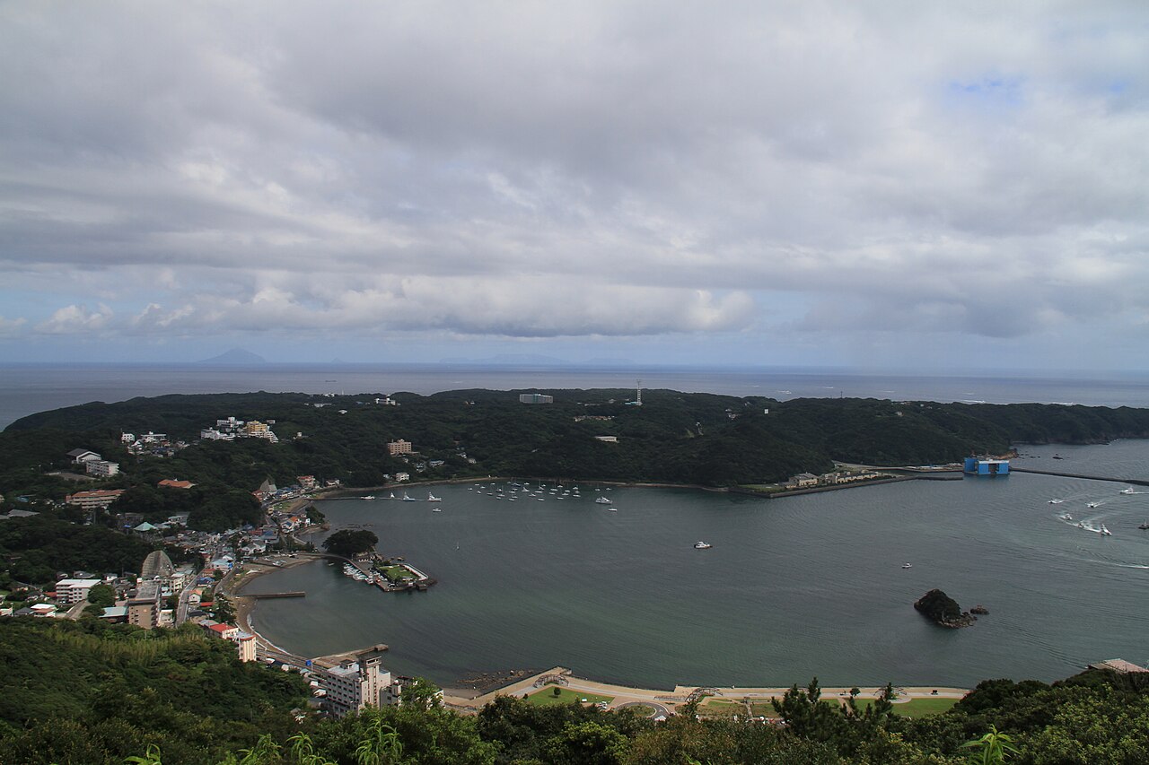 A calm bay ringed by wooded hills seen from a height, with small boats near the shore and a rocky islet in the water.