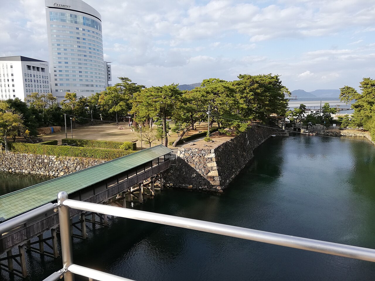 A covered wooden bridge crossing the inner moat at Tamamo Park (Takamatsu Castle ruins), with pines on the stone-walled bank and the sea beyond