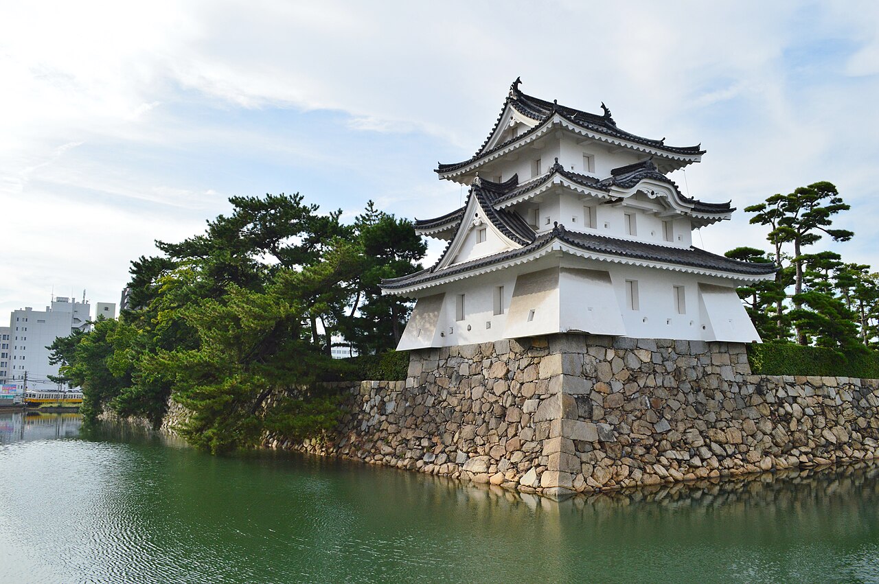 The white Ushitora Turret of Tamamo Park (Takamatsu Castle ruins) above a stone wall and green moat, with pine trees alongside