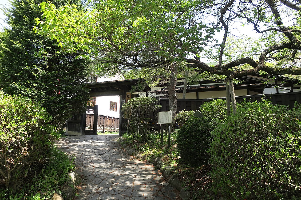 A flagstone path under leafy trees leading to the wooden gate and dark fence of the Ejima Enclosed Residence in Takato.
