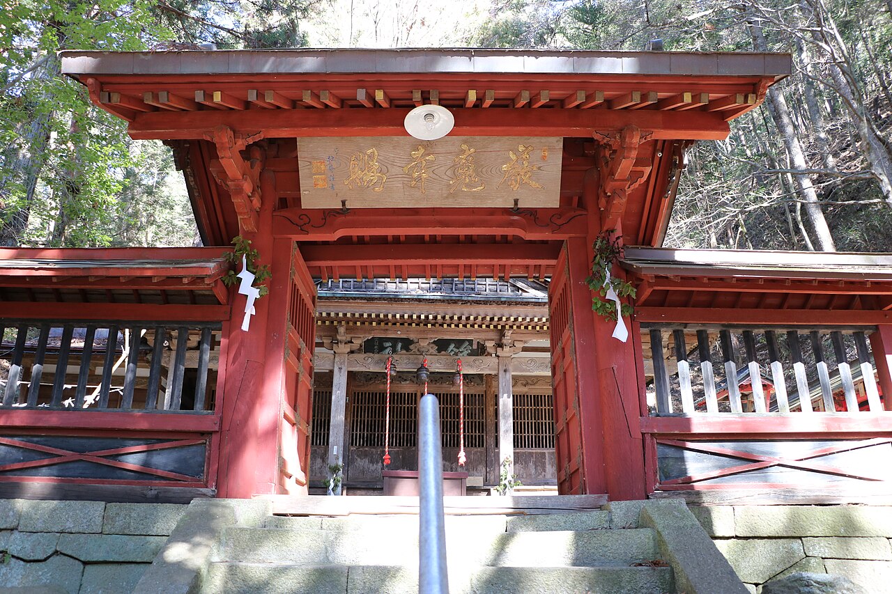 The vermilion gate of Hokomochi Shrine at the top of its stone steps, with a gilded wooden plaque under the eaves and bare trees behind.