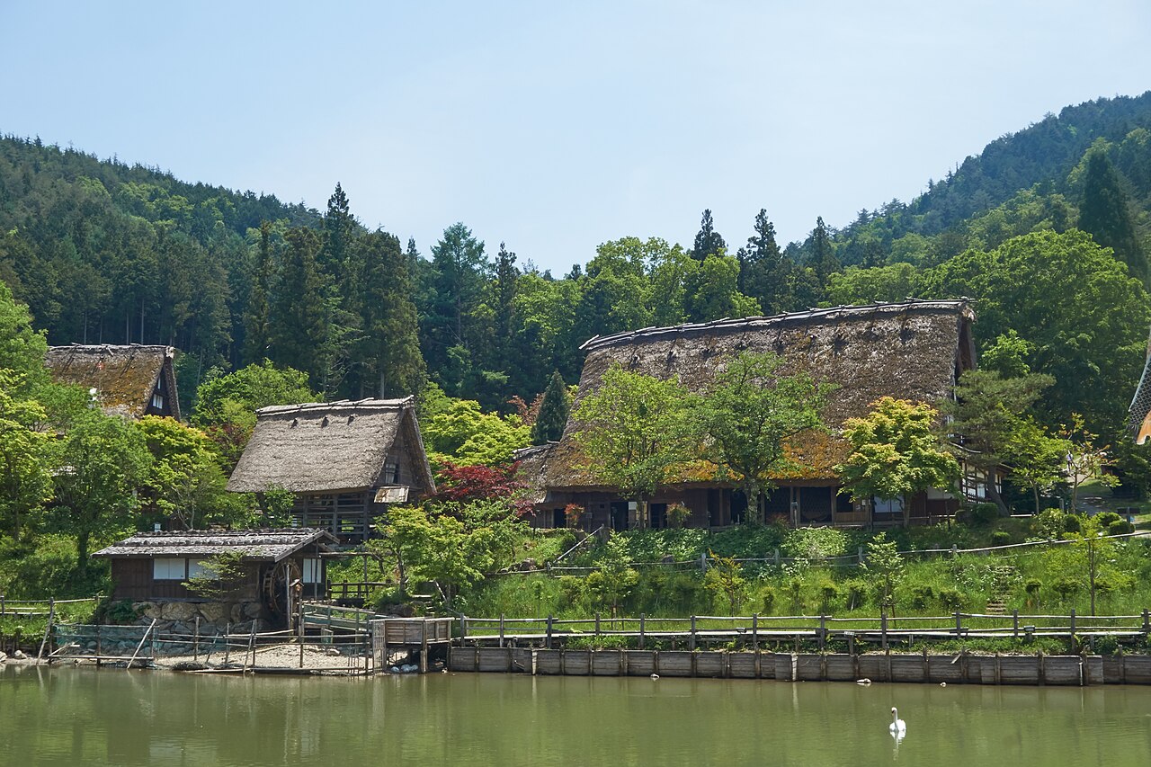 Thatched gassho-style farmhouses above a pond in early summer green at Hida no Sato open-air village in Takayama, with forested hills behind.